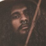 A striking portrait of a man with curly hair and hat in a natural outdoor setting.